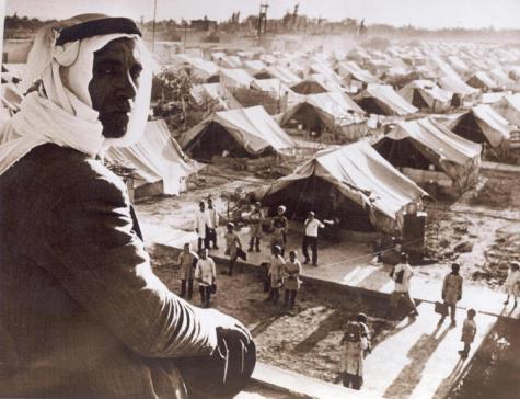 Black and white image of a Palestinian refugee camp after the mass displacement in 1948. Many white tents are lined up in the background and in the foreground there is a man turning to face the camera wearing a traditional head covering 