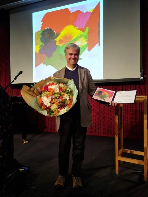 Image of World Beyond War co-founder David Swanson on stage receiving a prize holding a certificate and a large colourful bunch of flowers