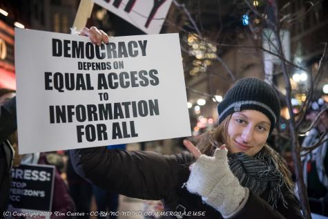 A smiling woman wearing dark winter clothes holds a sign which reads 'Democracy depends on equal access to information for all'
