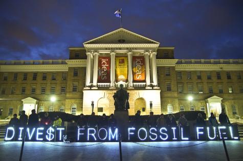Protesters stand infront of a governmental building at night with blue light up letters which read 'Divest from fossil fuel.'