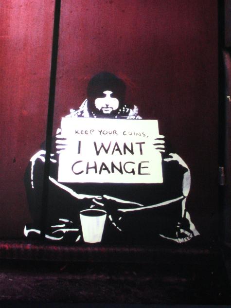 Photograph of street art. A man sits cross-legged on the floor holding a sign that says 'Keep your coins, I want change'