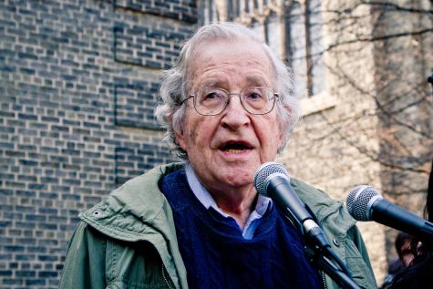 Noam Chomsky is an older white gentleman who is a celebrated linguist, and political analyst. He stands behind two microphones delivering a speech outside.