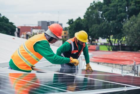Two men wearing green overalls, orange hi-vis jackets, and safety helmets are working on a installing a solar panel