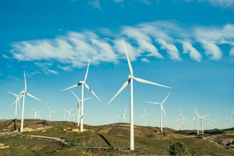 Many white wind turbines stand on a green hill under a blue and cloudy sky