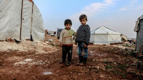 Two little boys stand on the dirty floor infront of many tents at a refugee campsite.