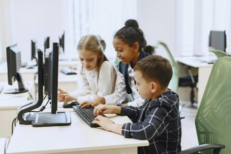 Three young students sit at a desk infront of a computer working. 