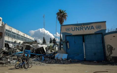 Large blue doors with the letters UNRWA painted above it with the United Nations symbol either side stand surrounded by rubble and destroyed buildings. Two men pushing bicycles are walking by.
