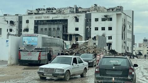 A destroyed hospital in Gaza has half of its front wall missing, it is surrounded by rubble, the whole scene is grey and dusty. There is an old bus and some cars parked infront of it.