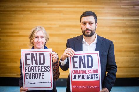 Photo to mark Migrants International Day. Two people hold up signs one which says 'End fortress Europe' and the other 'End Criminalisation of Migrants'.