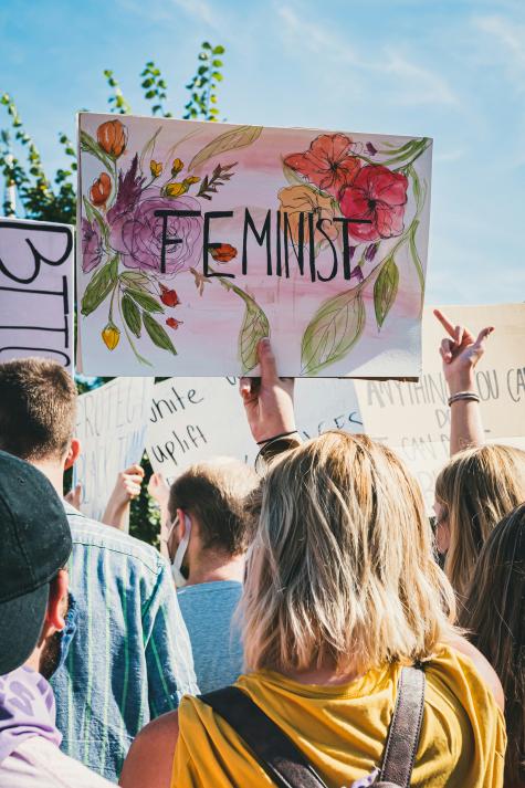 View from the back of a women's rights protest, there is a large white poster that says 'Feminist' with beautiful flowers painted in the background