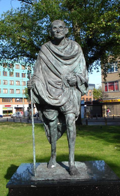 A statue of Gandi stands outside in the Netherlands. He is barefoot wearing traditional robes and holds a walking stick.