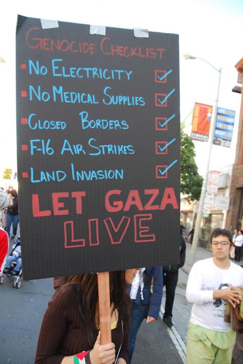 Gaza protest in San Francisco. A woman is holding a large black sign which has a gencoide checklist. Underneath is says 'Let Gaza Live.'