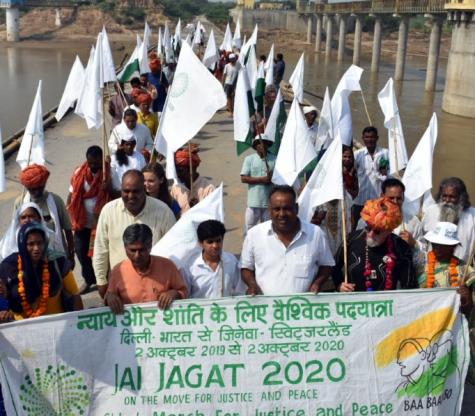 Jai Jagat 2020 crossing the Chambel river, Madhya Pradesh, India. Many people walk across a bridge holding white flags and a large banner with the organisations name on.