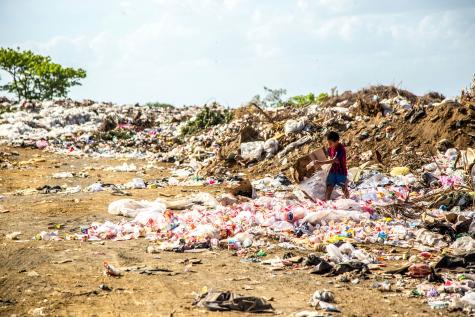A young boy is walking around a landfill site filled with plastic. He is carrying a cardboard box.