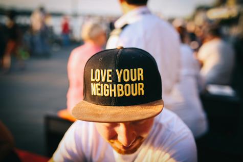 A man sits in a busy park wearing a cap that says 'Love Your Neighbour' 