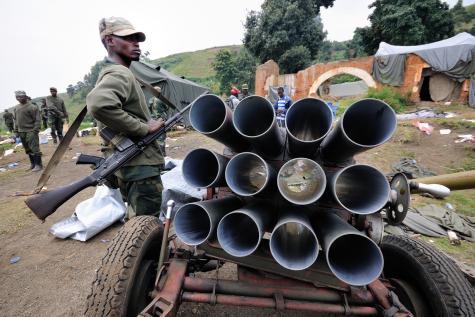 M23 Rebels in Congo. A man stands holding a gun next to a cart filled with metal pipes.