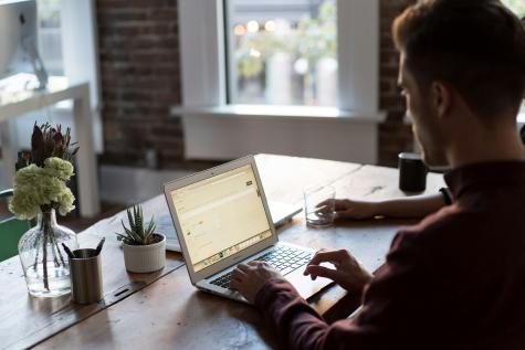 Image of a man from behind. He is working on his laptop at a wooden desk. 