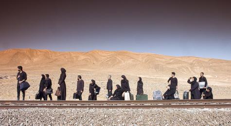 Many migrants line up and wait for a train with their luggage. They are in the dessert with no buildings around.