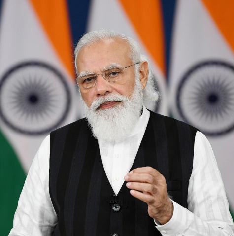 Narendra Modi, Prime Minister of India. An older gentleman stands infront of the Indian flag delivering a speech, he is smartly dressed in a white shirt and blue waistcoat. He has a large grey beard and wears glasses.