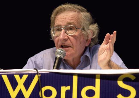 Noam Chomsky sits delivering a speech with a microphone behind a podium at the World Social Forum in 2003.