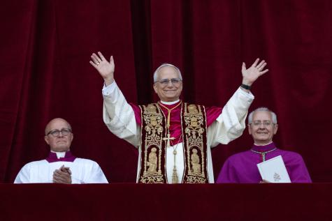 Pope Leo XIV addresses an audience with open hands, he is wearing white and purple robes and is smiling warmly.