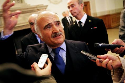 Image of Prince of Jordan Hassan bin Talal. Wearing a dark suit he is surrounded by reporters as he holds his hand in the air.