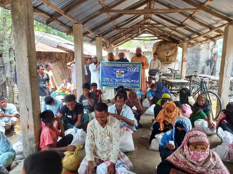 Many people sit and eat on the floor under a wodden structure. Four men stand at the back holding a sign which says 'Ramadan Food Distribution 2022 - Needy Foundation'