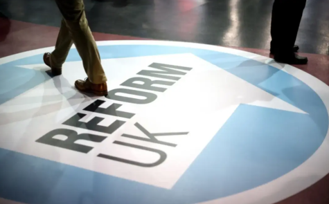 The blue symbol of British political party Reform UK is displayed on the floor of a large room, there are people legs and feet shown in the image as the walk over the sign.