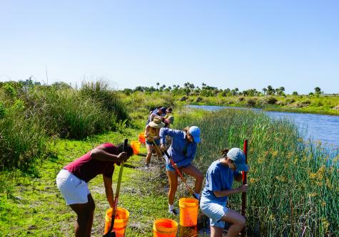Seven volunteers work under the sun next to a river, they are holding shovels and each have an orange bucket.