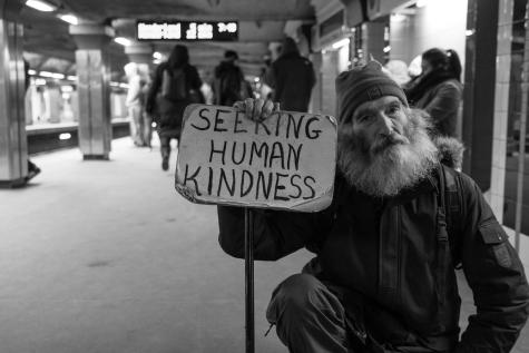 Black and white photograph of man holding up a cardboard sign that says 'Seeking human kindness.' He is sitting on the floor of a subway station, he has a large beard and is wearing a beanie. 