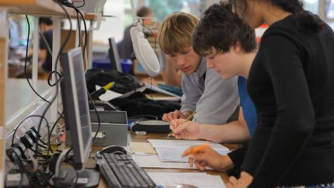Three teenage students are concentrating in a science class. They are infront of a computer looking at notepads.