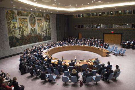 Photo taken inside a Security Council meeting where members all sit around a large circular desk. There are enormous ornate paintings covering one side of the wall. 