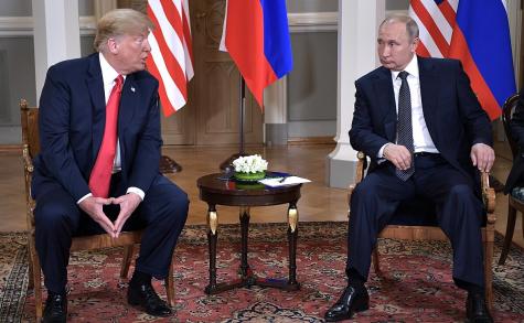 Vladimir Putin & Donald Trump at a meeting in Helsinki. They sit next to a small table with their respective flags in the background.