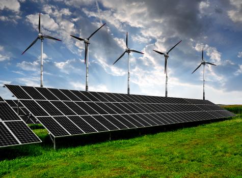 A large row of blue solar panels sits on a green field with tall white wind turbines in the background