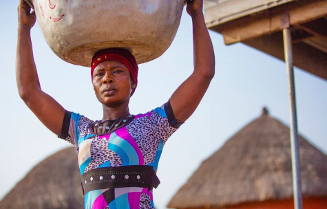 A women from Ghana carries a large metal bowl on her head, she wears a colourful dress and her expression is serious. 