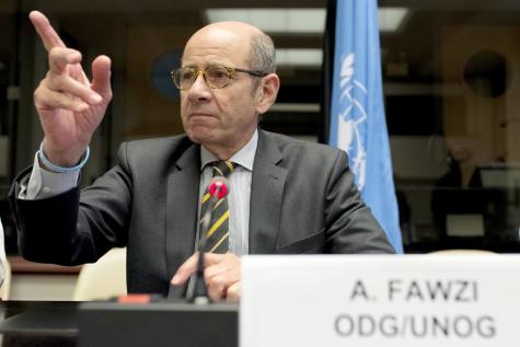 Ahmad Fawzi sits at a United Nations meeting wearing a great suit and glasses, he has his finger raises in the air.