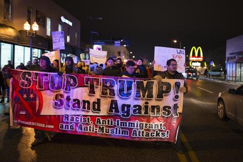 A diverse group of protesters holds a large banner reading "STOP TRUMP! Stand Against! Racist, Anti-Immigrant, and Anti-Muslim Attacks!" at night.