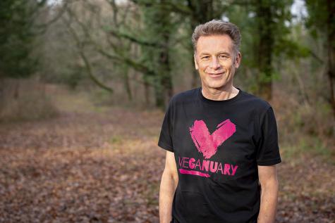 Image of Chris Packham the British naturalist standing in a beautiful forest. He wears a black Veganuary tshirt.