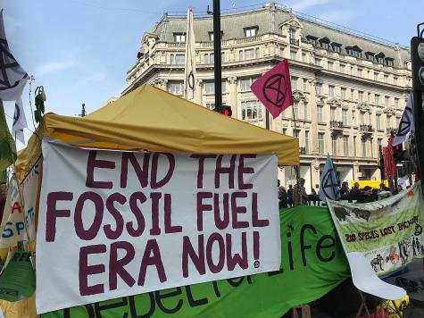 A yellow tent in an outdoor protest scene displays a large banner that reads "END THE FOSSIL FUEL ERA NOW!" with various flags and signs around.