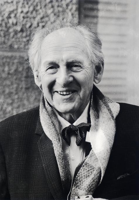 Foto von Leopold Kohr in 1983 - A smiling elderly man with light hair, wearing a dark suit and scarf, stands against a textured wall.