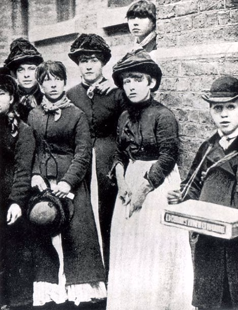 A historical black-and-white photograph of a group of eight young people, primarily women in long dresses and hats, standing together outdoors.