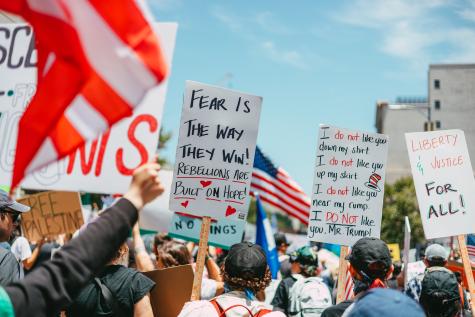 A crowd of protesters holds signs with messages about fear and oppression, surrounded by flags and a clear blue sky.