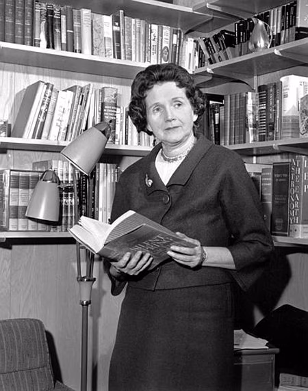 A woman stands in a library, holding a book. She wears a dark suit with a pearl necklace, surrounded by shelves of books.