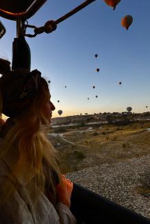 A silhouette of a women from a hot air balloon looks out into the sunrise. There are many other balloons in the sky in the background. 