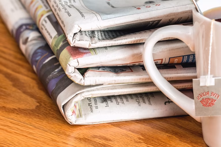Cup of tea sits on a wooden table next to a stack of folded newspapers