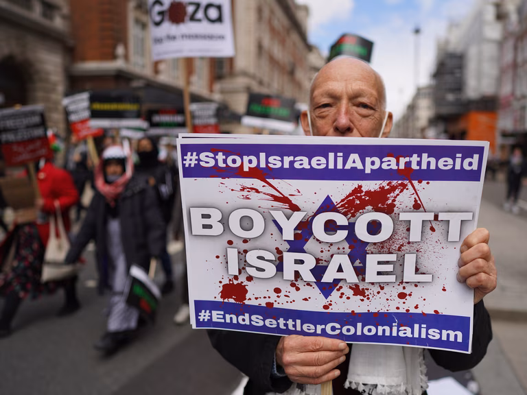 An older white gentleman stands in the street holding a large white sign which says ' Boycott Israel' the man is part of a larger protest which we can see part of in the background