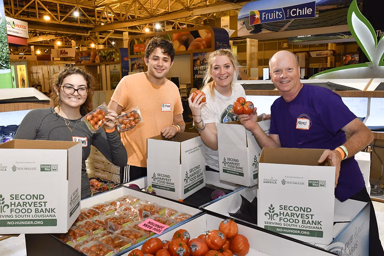 Two men and two women happily stand behind boxes of donated food in a food bank. They re wearing name tags and are holding fresh tomatos 