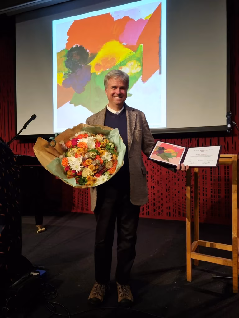 Image of World Beyond War co-founder David Swanson on stage receiving a prize holding a certificate and a large colourful bunch of flowers 