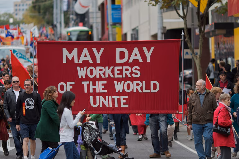 May Day March in Melbourne, Australia. Protesters hold up a large red sign that says 'Workers of the World Unite.'