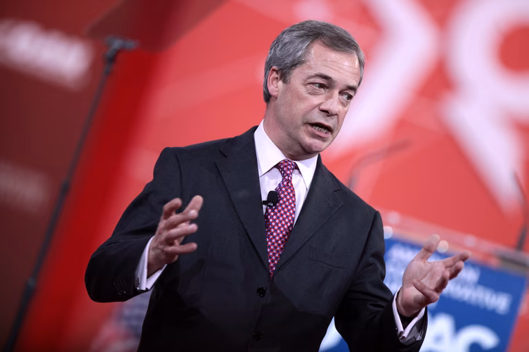 Nigel Farage speaking at the 2015 Conservative Political Action Conference. He stands infront of a red background wearing a black suit holding his hands open and looking away from the camera